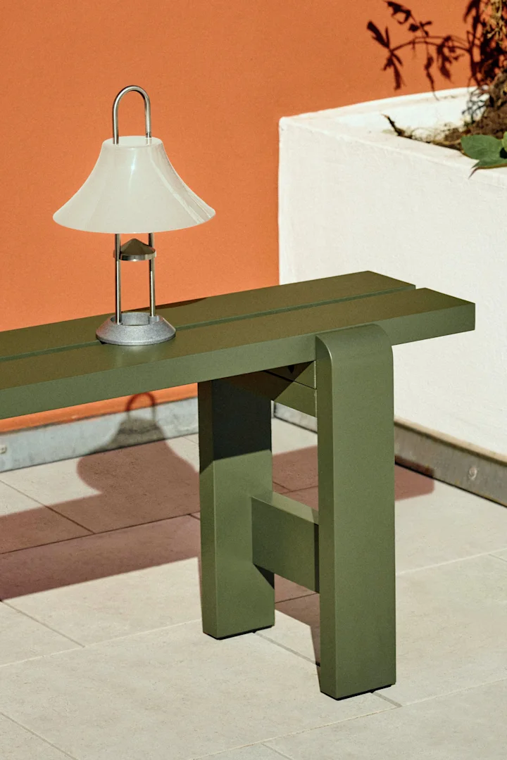 A modern table lamp with a frosted white shade on a dark green metal bench against an orange wall.
