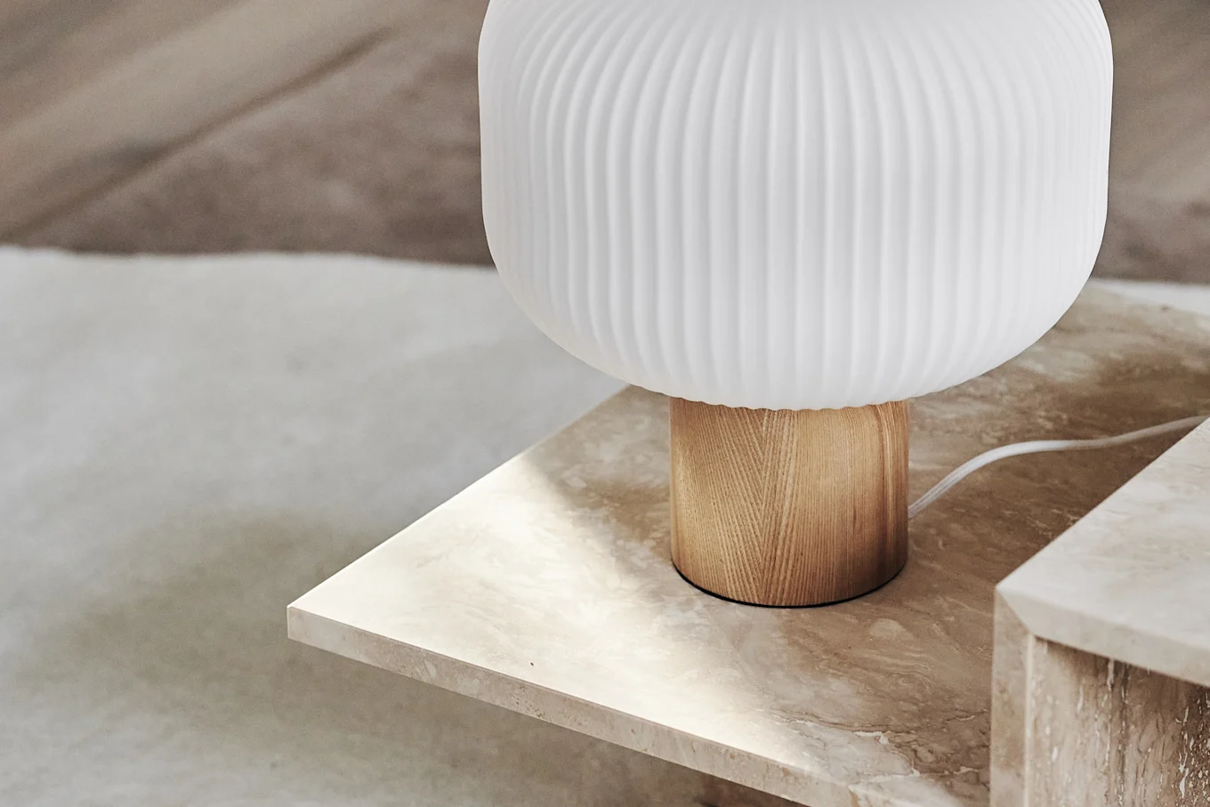 A close-up of a modern table lamp with a ribbed white glass shade and light wooden base on a beige marble table.
