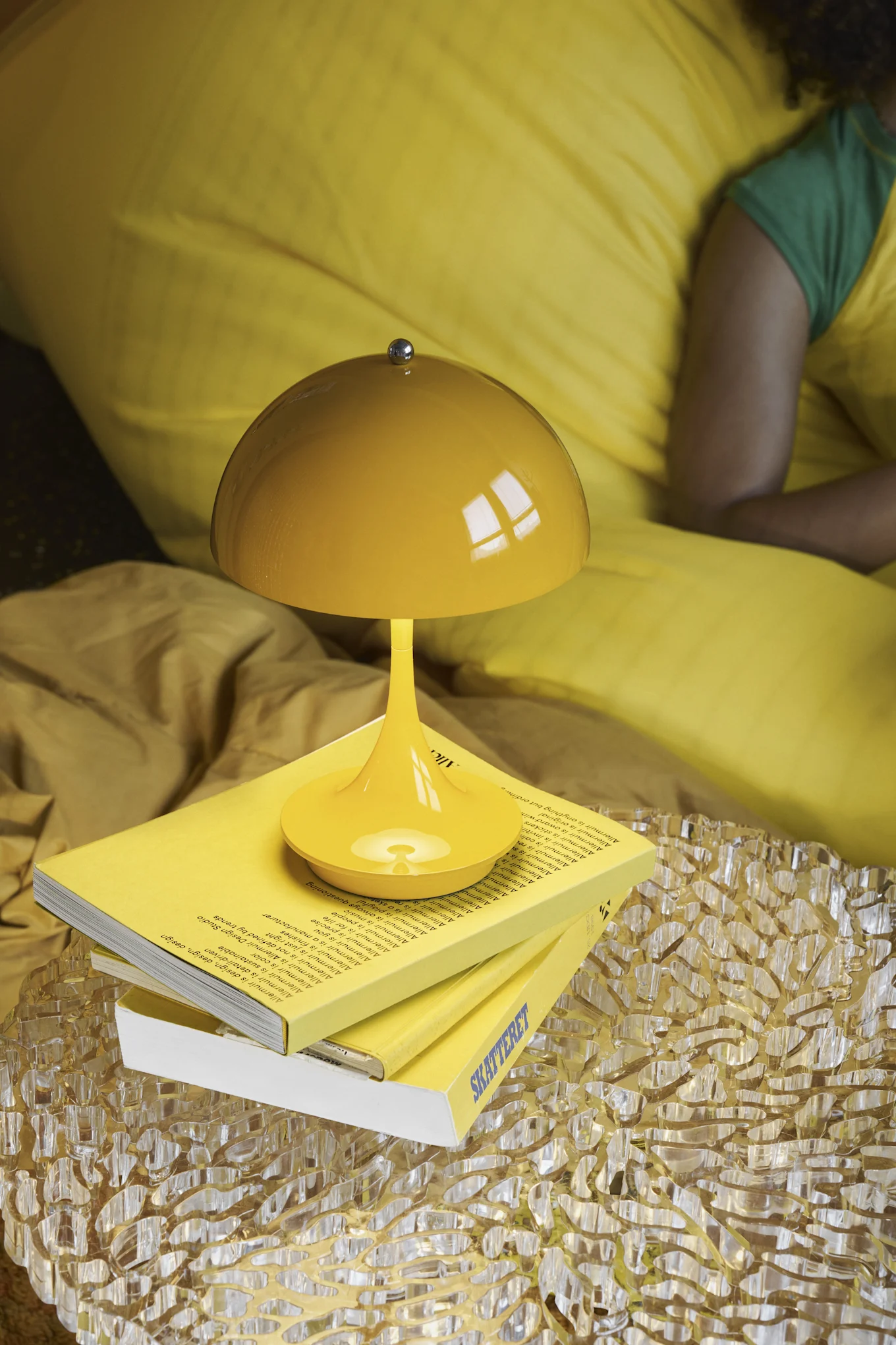 Yellow mushroom lamp on yellow books on a textured table, with yellow bedding and a person lying in the background.