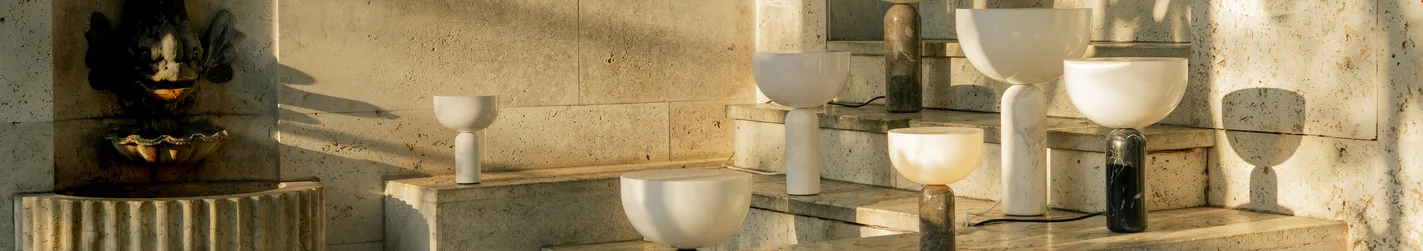 Modern lamps with bowl-shaped tops and cylindrical marble bases on sunlit stone steps next to a fountain.