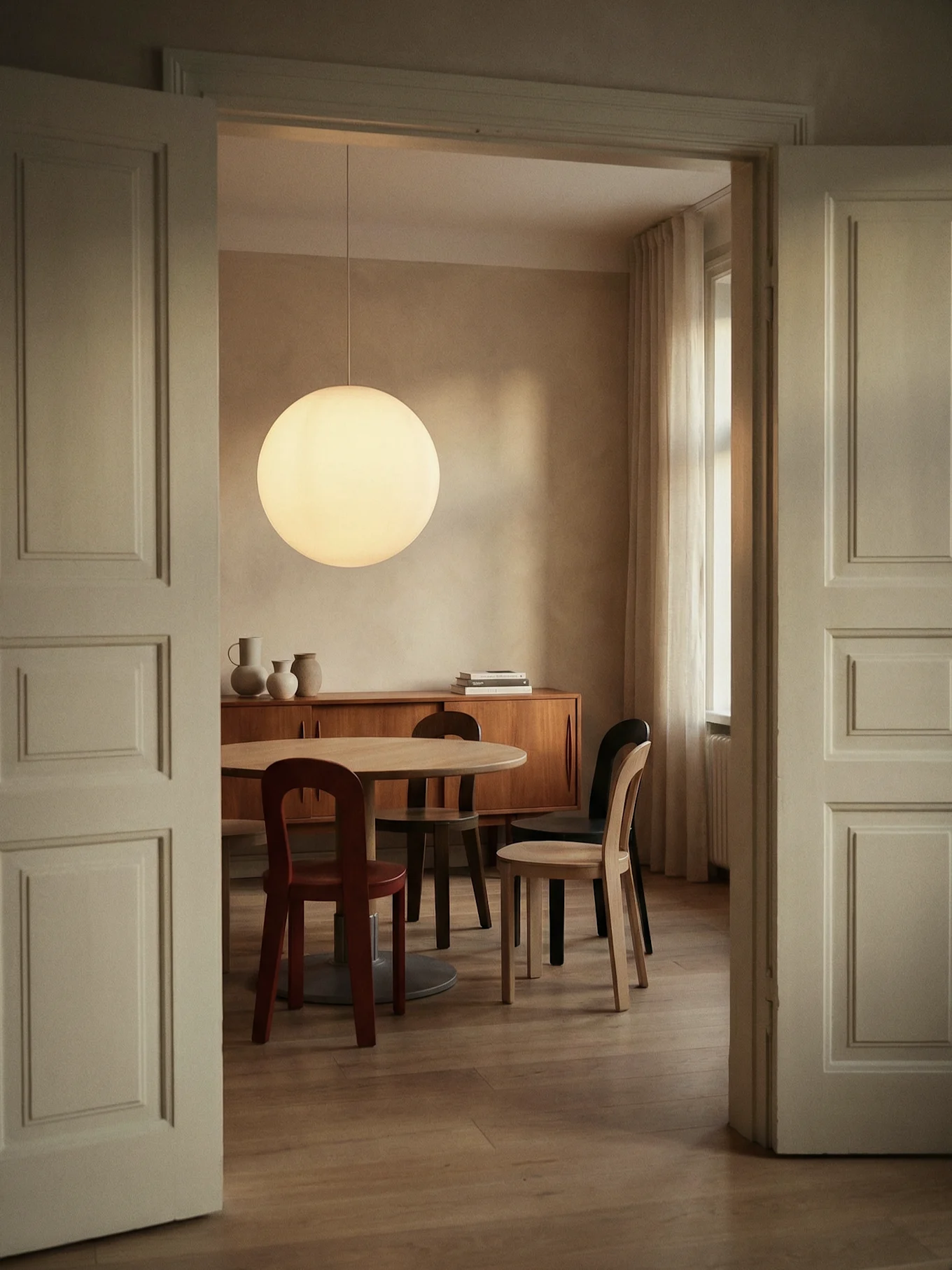 Dining room with a round wooden table, various chairs, a large spherical pendant lamp, and a wooden sideboard, seen through an open doorway.