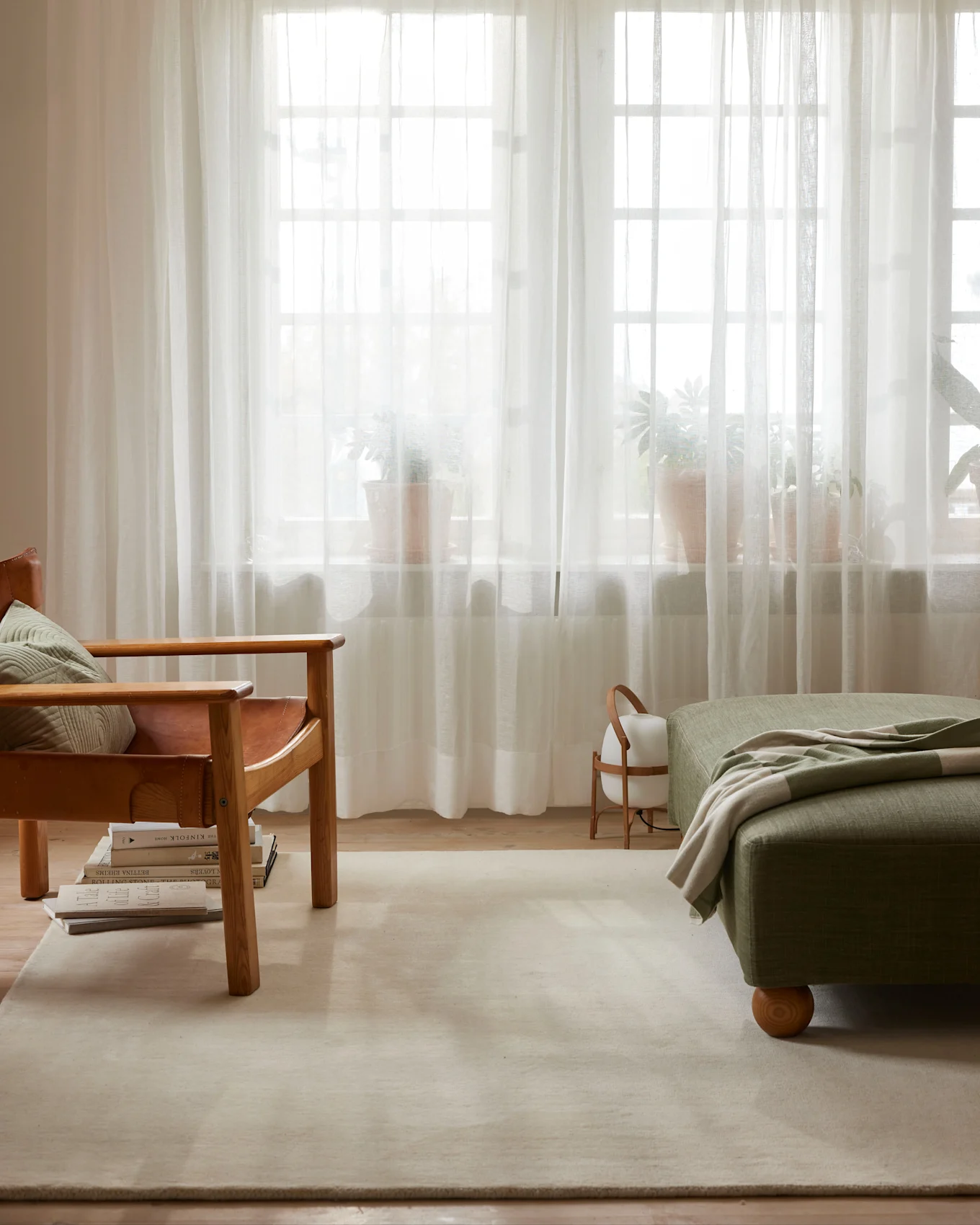 Living room with a brown leather armchair, green ottoman with a striped blanket, sheer curtains, and potted plants.
